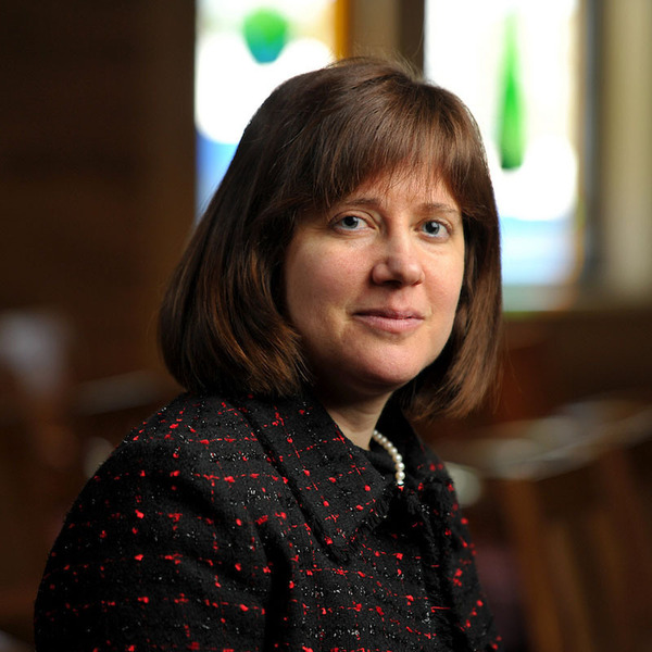 Smiling woman with brown hair and blue eyes, wearing a black tweed jacket with red flecks and a pearl necklace. Blurred stained glass.