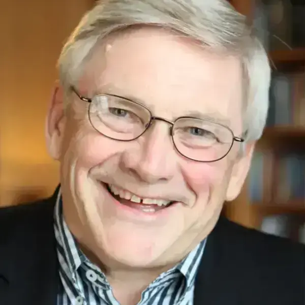 A man smiles warmly, wearing glasses, a striped shirt, and a dark jacket, with bookshelves behind him.