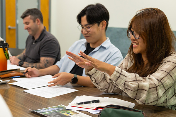Smiling woman in plaid shirt gestures while discussing with two engaged men in a classroom.